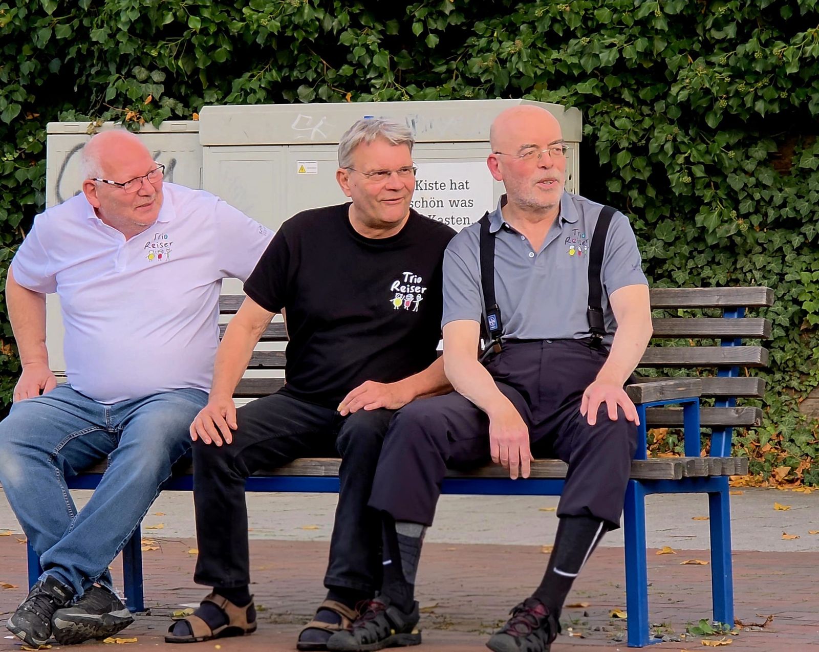 Die drei Musiker von Trio Reiser sitzen in Bandshirts nebeneinander auf einer Parkbank